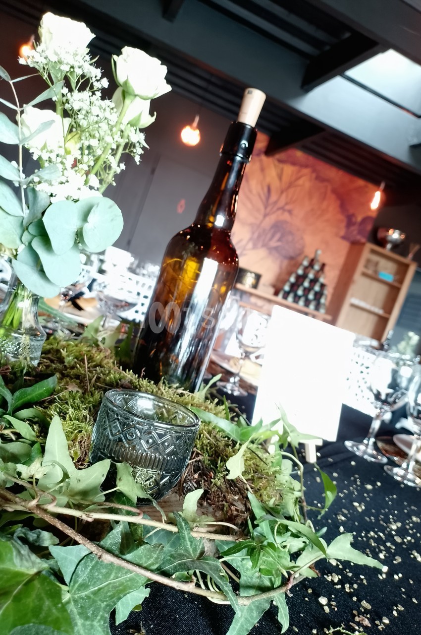 Centre de table avec une bouteille en verre, feuillage, mousse et bougie, dans une salle décorée pour un événement.