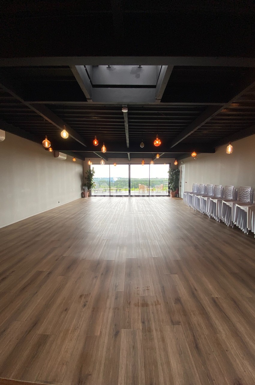 Salle vide avec parquet en bois, éclairée par des suspensions lumineuses, donnant sur une baie vitrée avec vue extérieure.