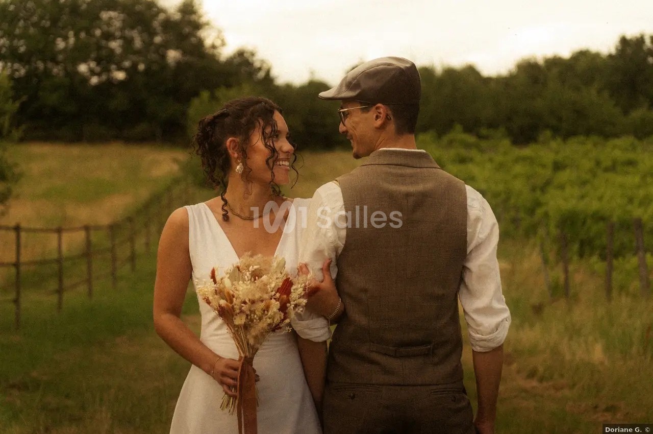 Un couple en tenue de mariage se tient dans un vignoble, la mariée tenant un bouquet de fleurs séchées.