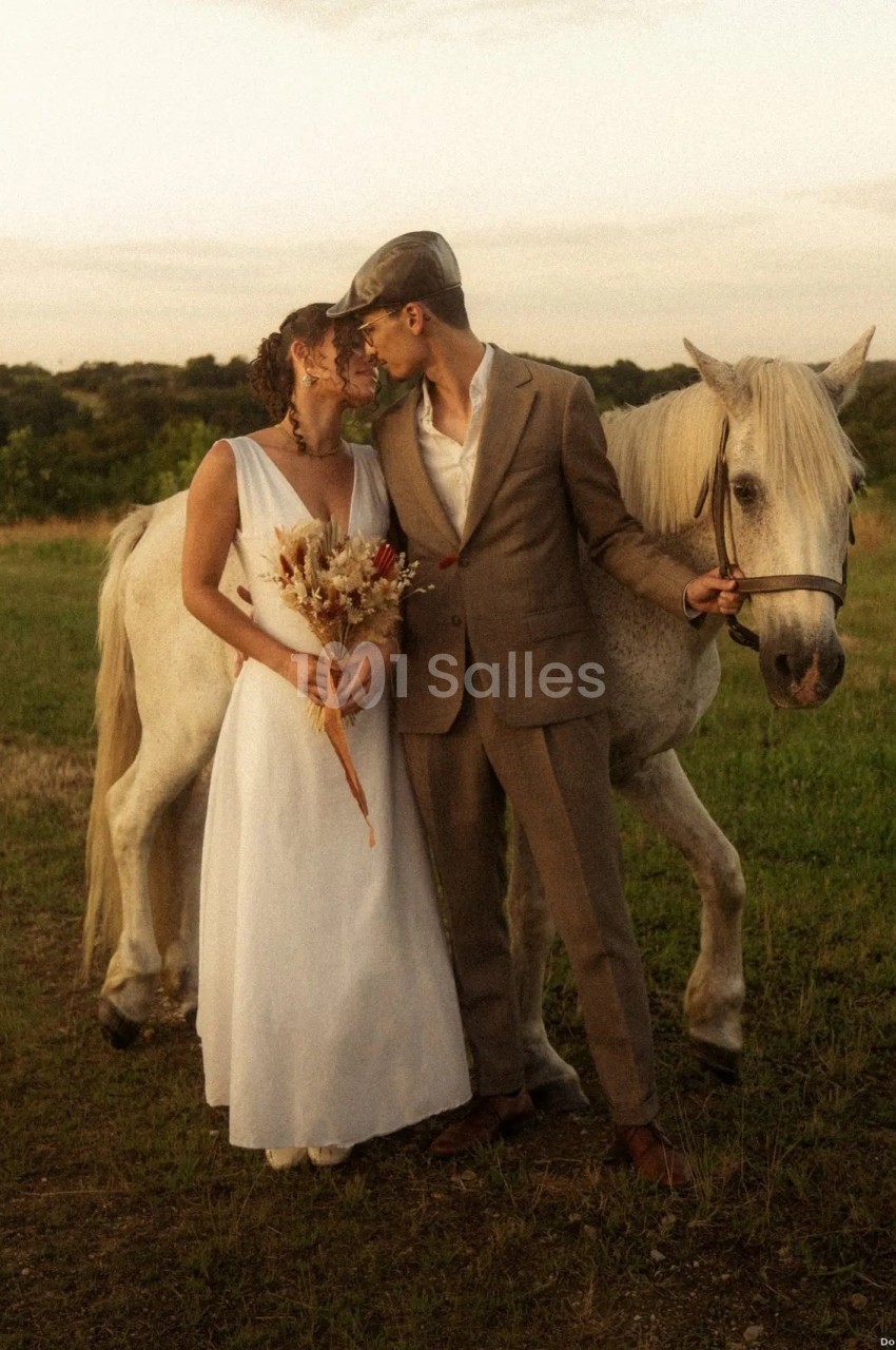 Un couple en tenue de mariage pose près d'un cheval blanc dans un champ au coucher du soleil.