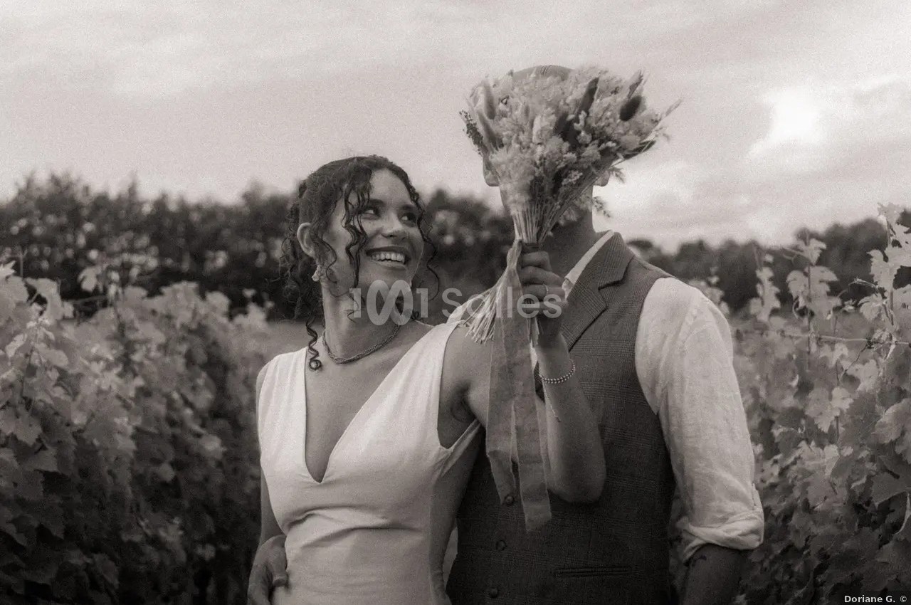 Un couple dans un vignoble, la femme sourit tandis que l'homme cache son visage avec un bouquet de fleurs.