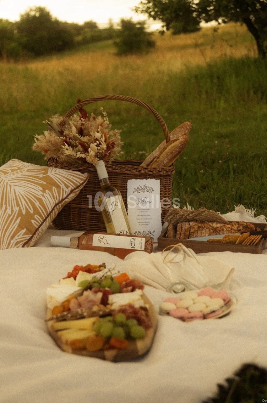 Pique-nique champêtre avec panier en osier, pain, fruits, fromage, macarons et bouteille de vin sur une nappe blanche.