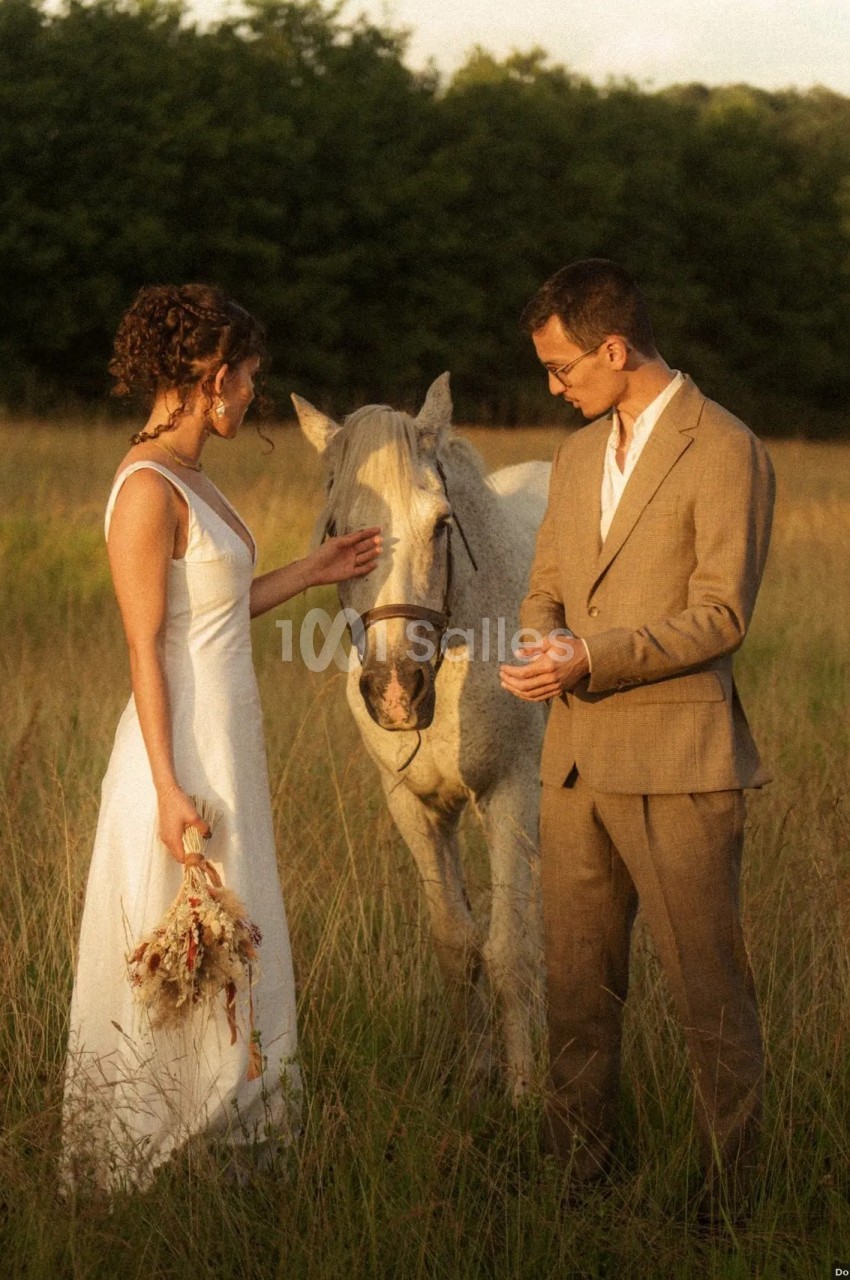 Une femme en robe blanche et un homme en costume beige se tiennent près d'un cheval dans un champ herbeux.