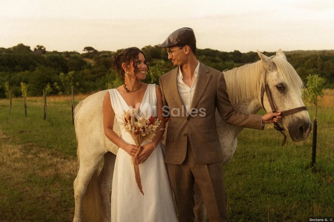 Un couple souriant en tenue élégante se tient près d'un cheval blanc dans un champ verdoyant au coucher du soleil.