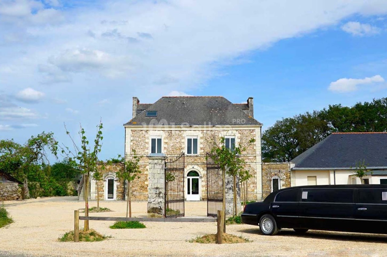 Façade d'une maison en pierre avec portail en fer forgé, entourée d'arbres, et une limousine noire stationnée devant.