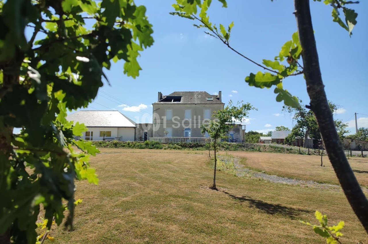 Grande maison en pierre avec toit mansardé, entourée d'un vaste jardin herbeux, vue à travers des branches d'arbres.