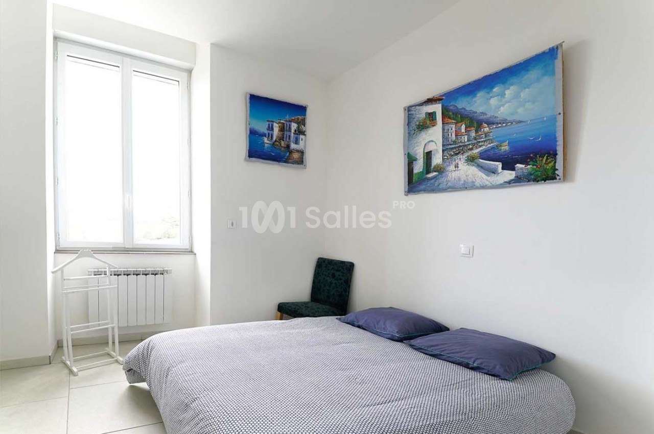 Chambre lumineuse avec un lit double, une chaise, un petit bureau blanc et des tableaux représentant des paysages côtiers.