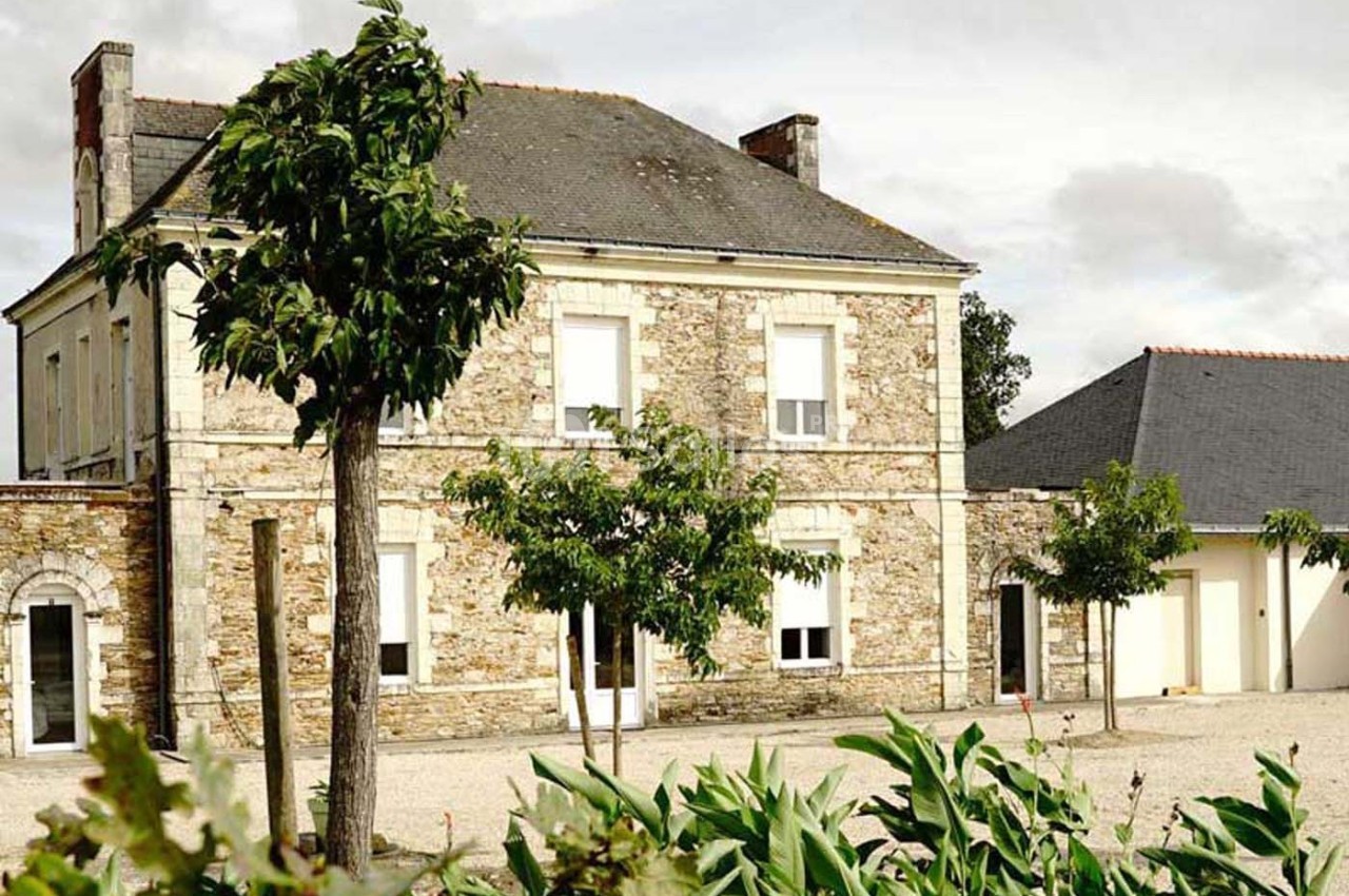 Maison en pierre avec toit en ardoise, entourée d'arbres et de végétation, sous un ciel partiellement nuageux.