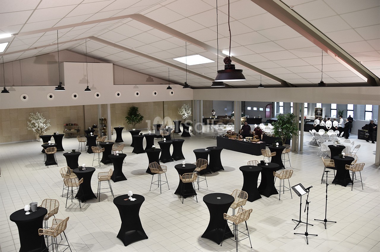 Salle de réception aménagée avec des tables hautes noires, des chaises en rotin et un buffet en arrière-plan.