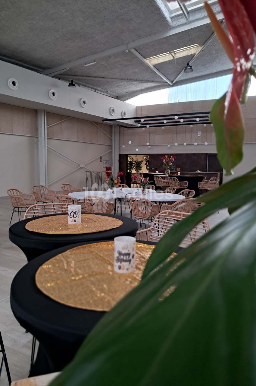 Salle de réception avec tables décorées, nappes dorées et chaises en rotin sous un plafond avec puits de lumière.