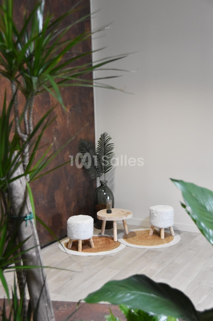 Deux tabourets blancs et une petite table en bois sur des tapis ronds, entourés de plantes dans un espace lumineux.
