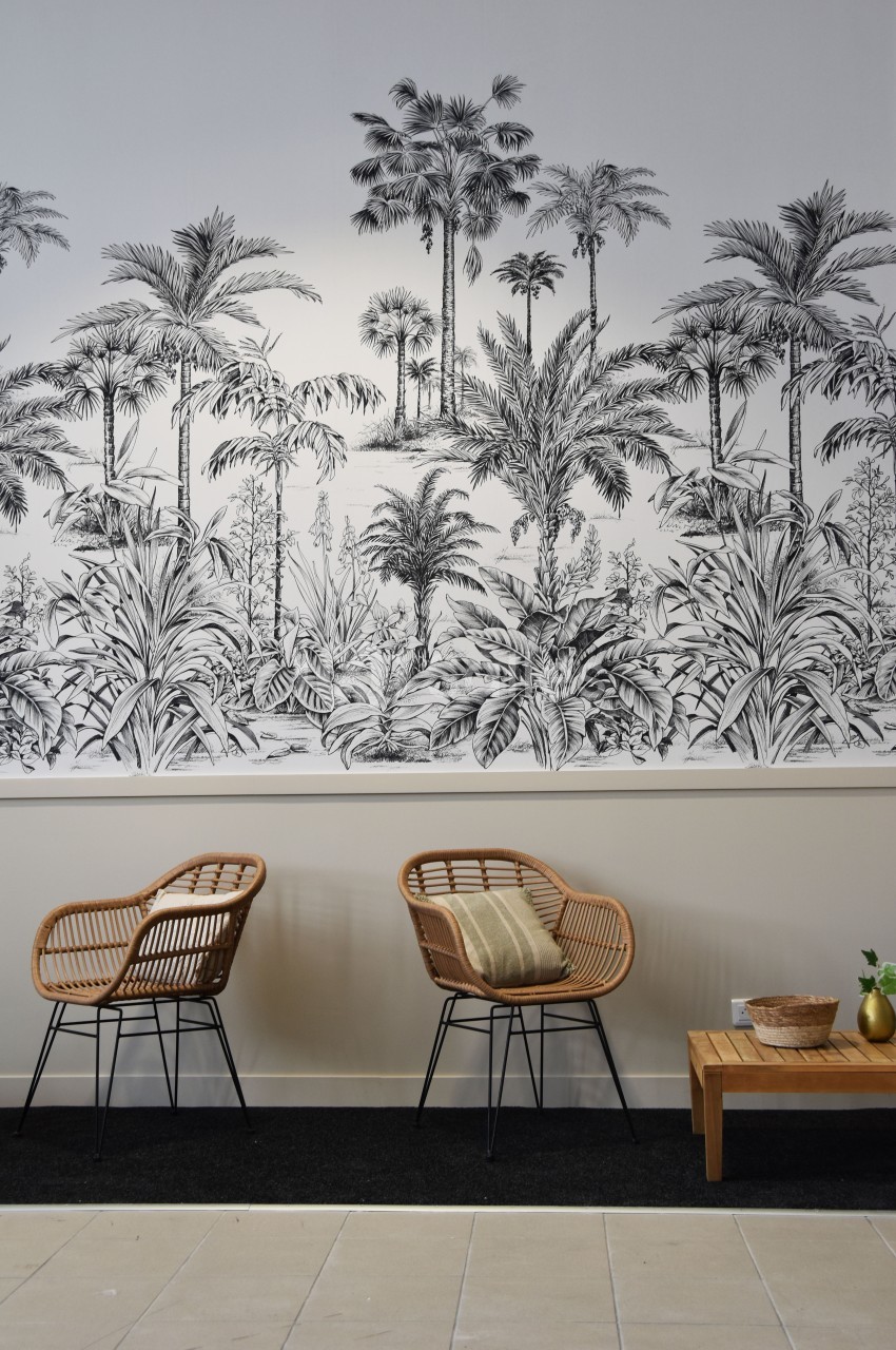Deux fauteuils en rotin et une table basse en bois devant un mur décoré d'une fresque de palmiers en noir et blanc.