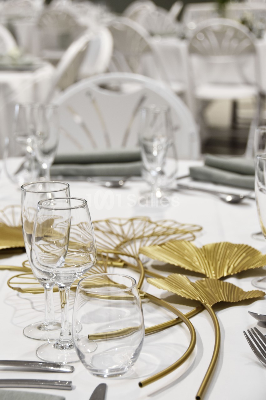 Table dressée avec nappes blanches, verres, couverts et décoration dorée en forme de feuilles.