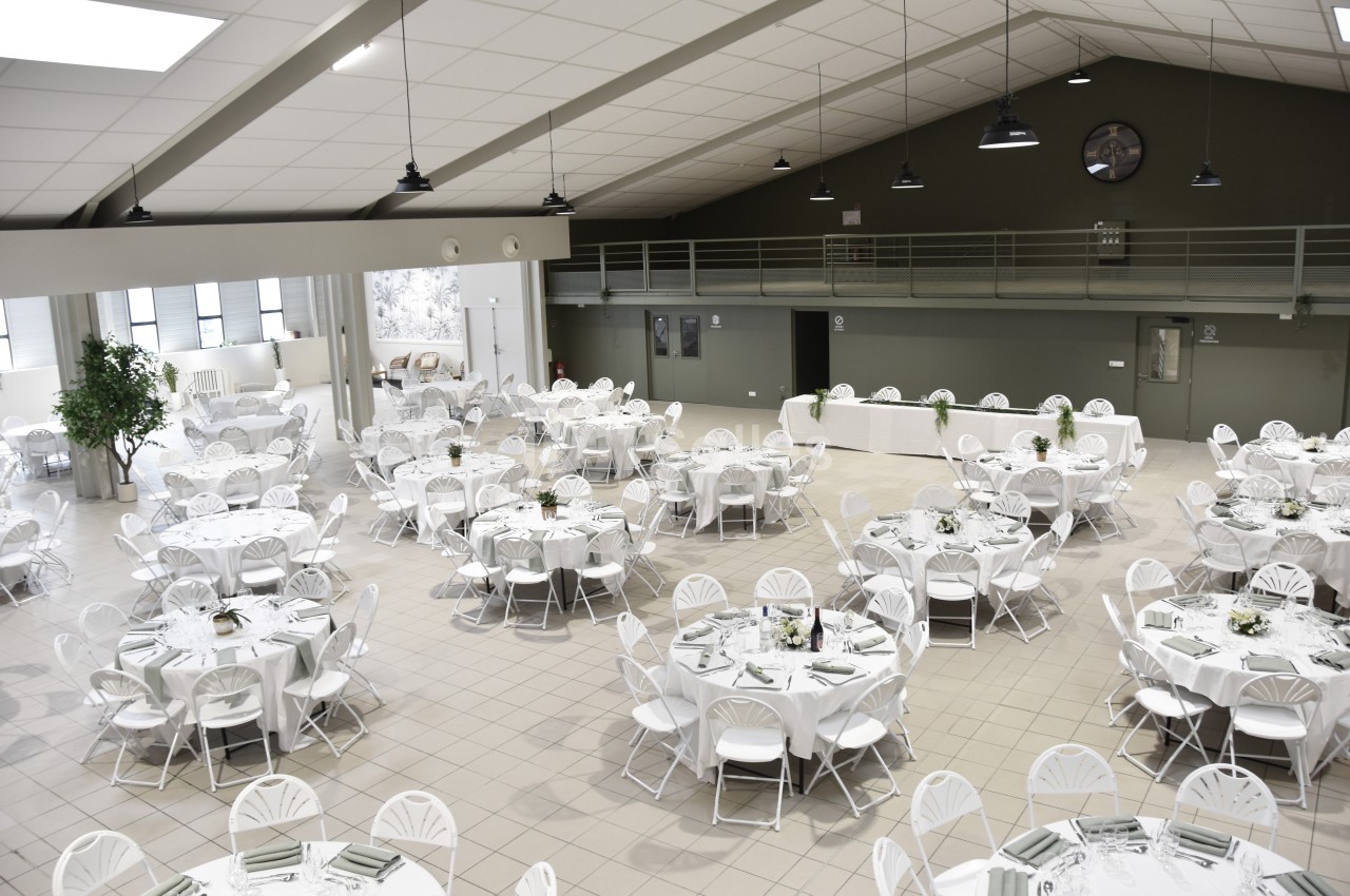 Salle de réception spacieuse avec tables rondes dressées et chaises blanches, éclairée par des suspensions noires.