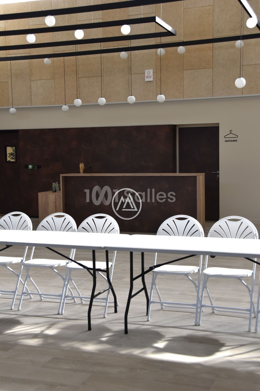 Salle lumineuse avec un comptoir en bois foncé, logo circulaire blanc, table et chaises blanches au premier plan.