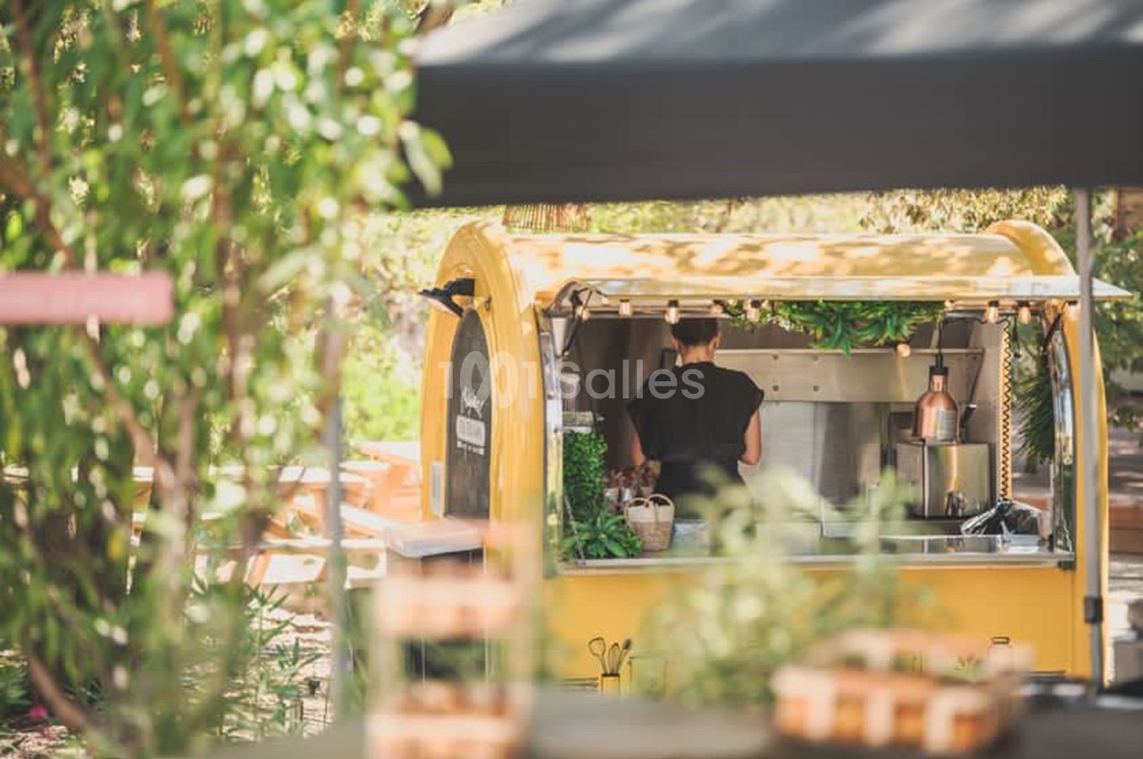 Un food truck jaune dans un cadre extérieur verdoyant, avec une personne préparant des plats à l'intérieur.