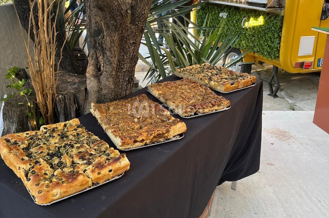Quatre focaccias variées disposées sur une table noire en extérieur, près d'arbres et d'une remorque jaune.