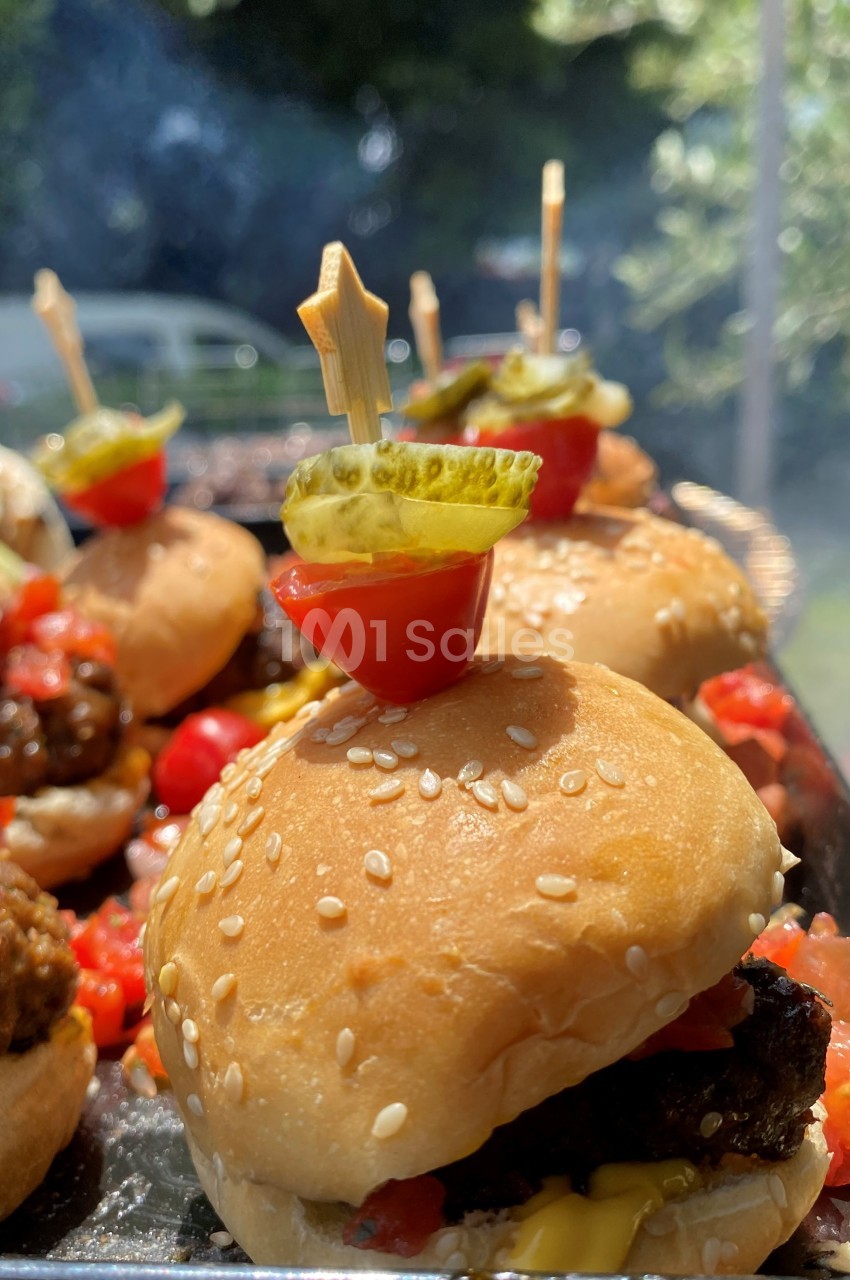 Mini burger garni de cornichon et tomate cerise sur un pique, avec d'autres burgers en arrière-plan.