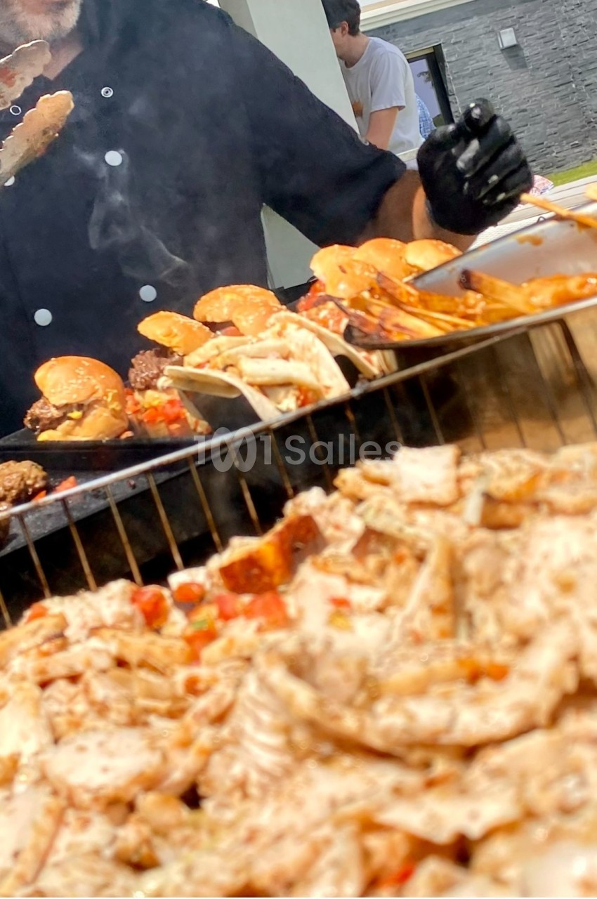 Chef cuisinant des viandes grillées et des burgers sur un barbecue en plein air, avec des convives en arrière-plan.
