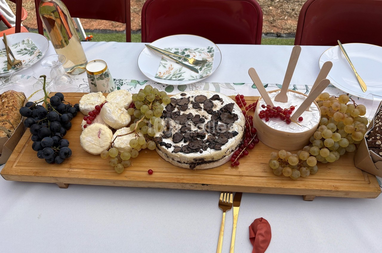 Plateau en bois avec fromages, raisins, truffes et groseilles, disposé sur une table dressée en extérieur.