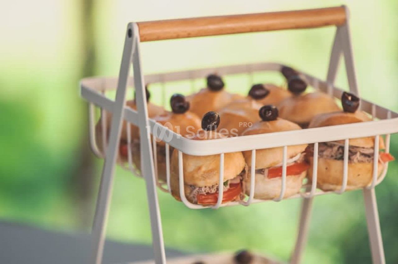 Plateau en métal contenant des mini-burgers garnis de tomates, olives noires et viande, posé en extérieur.