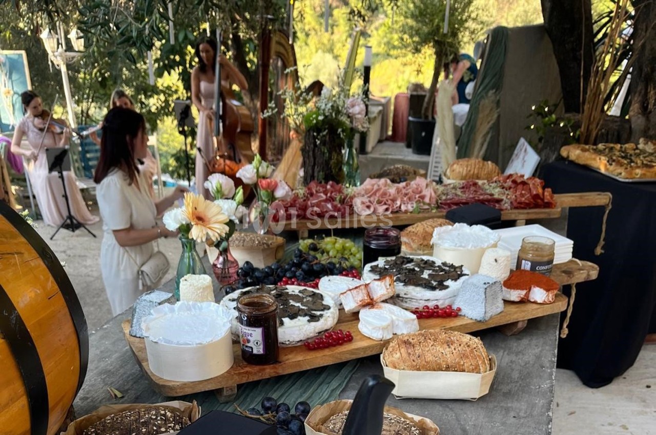 Plateau de fromages, charcuteries et pains sur une table en extérieur, avec des musiciens en arrière-plan.