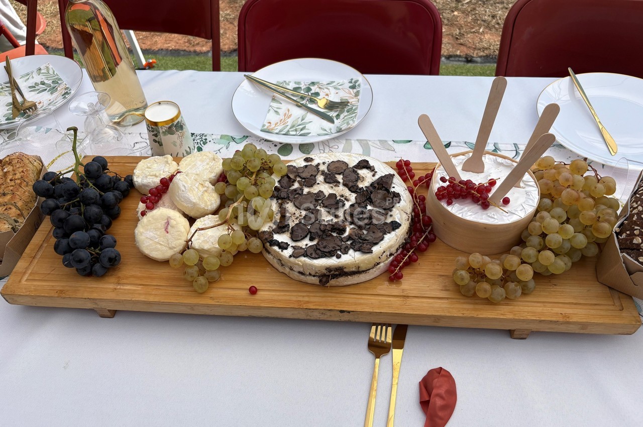 Plateau en bois avec fromages, raisins, truffes et groseilles, disposé sur une table dressée en extérieur.