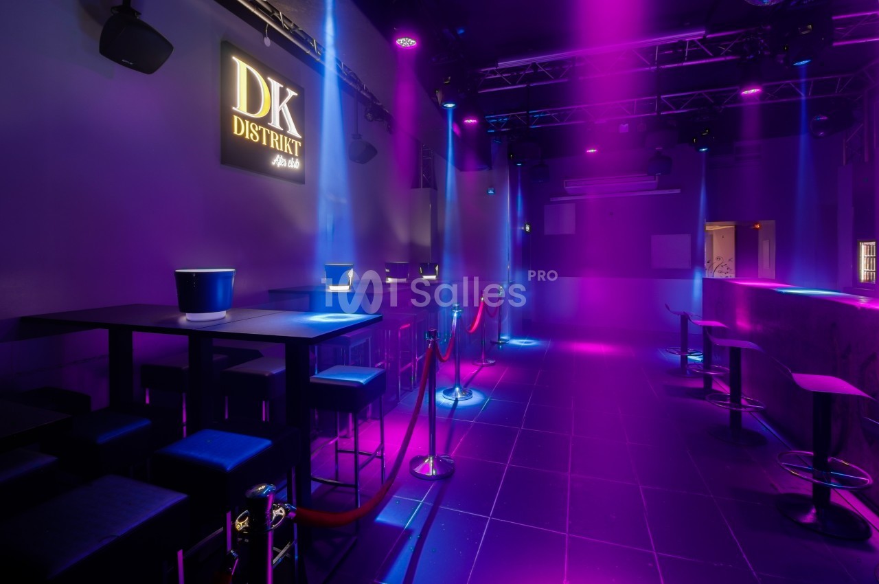 Salle de club avec éclairage violet et bleu, tables hautes, tabourets, cordons de séparation et logo mural lumineux.