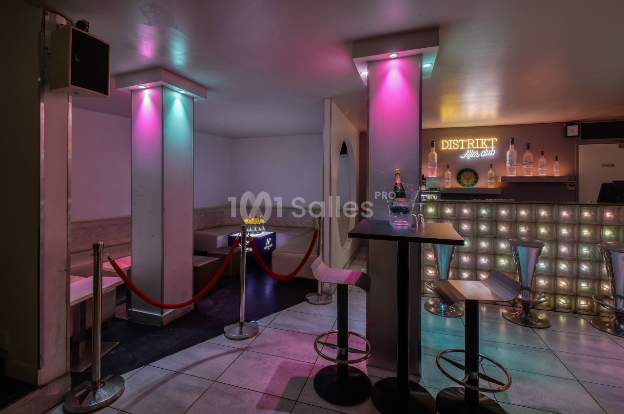 Intérieur d'un bar avec comptoir illuminé, tabourets, table haute et espace VIP délimité par des cordons rouges.