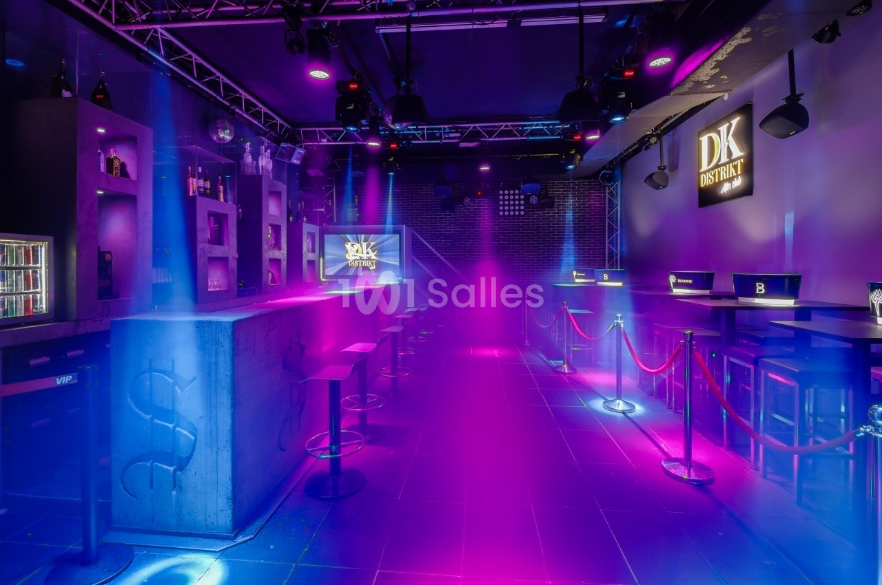 Salle de bar moderne avec éclairage coloré, tables hautes, tabourets et cordons de séparation.