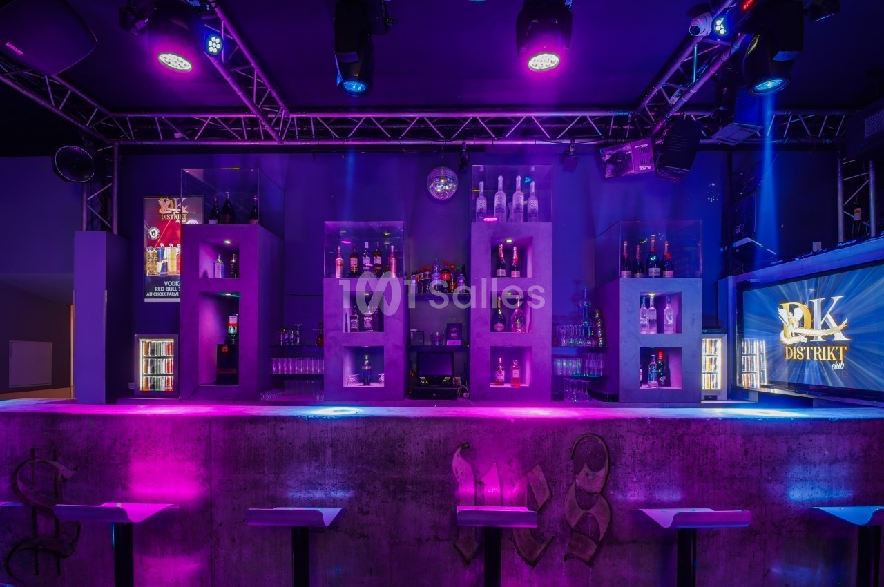 Bar moderne avec étagères rétroéclairées exposant des bouteilles, éclairage violet et ambiance nocturne.