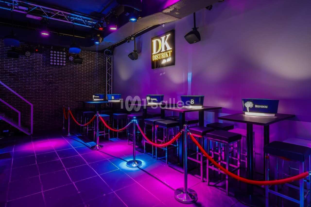 Salle de bar avec tables hautes, tabourets, éclairage coloré et cordons rouges délimitant les espaces.