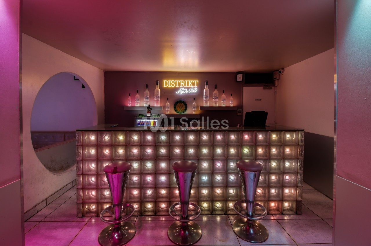 Bar avec comptoir en briques de verre illuminées, trois tabourets hauts et enseigne lumineuse ’Distrikt’ en arrière-plan.