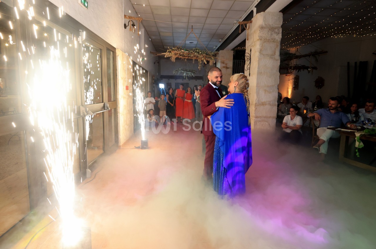 Un couple danse au milieu de fumée colorée et d'étincelles dans une salle décorée, entouré d'invités.