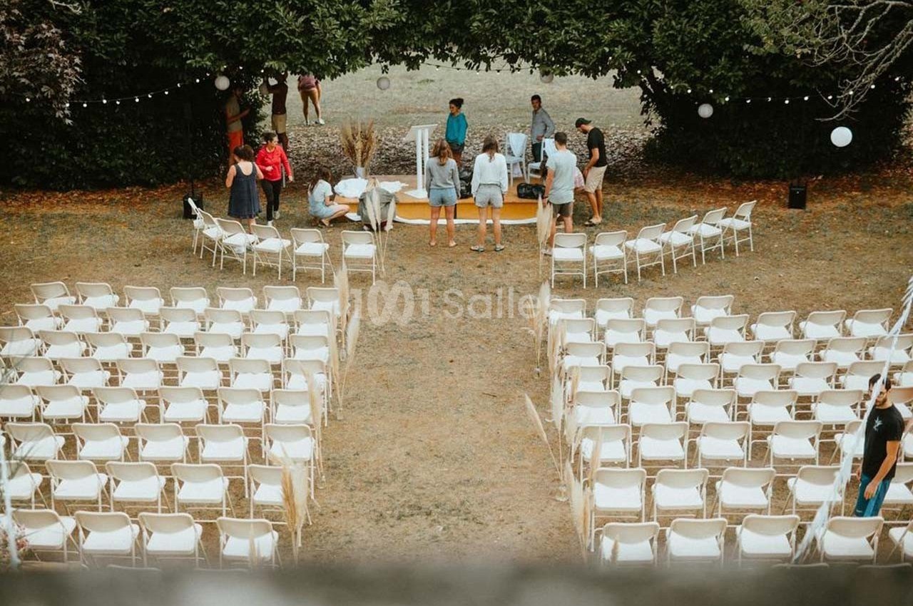 Chaises blanches disposées en rangées en extérieur, avec quelques personnes préparant une scène sous des arbres.