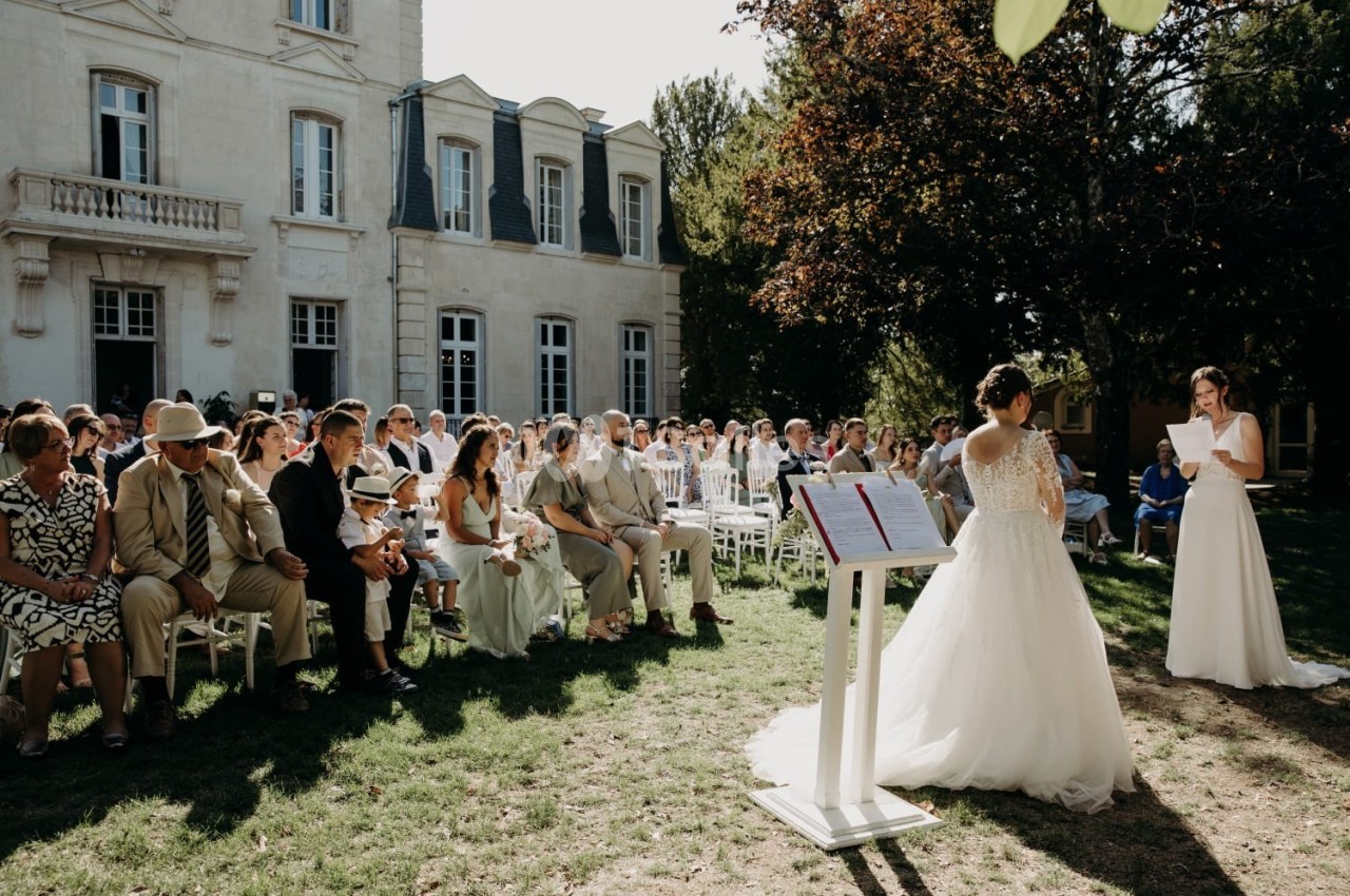 Une cérémonie de mariage en extérieur devant un bâtiment ancien, avec des invités assis et deux mariées échangeant leurs…