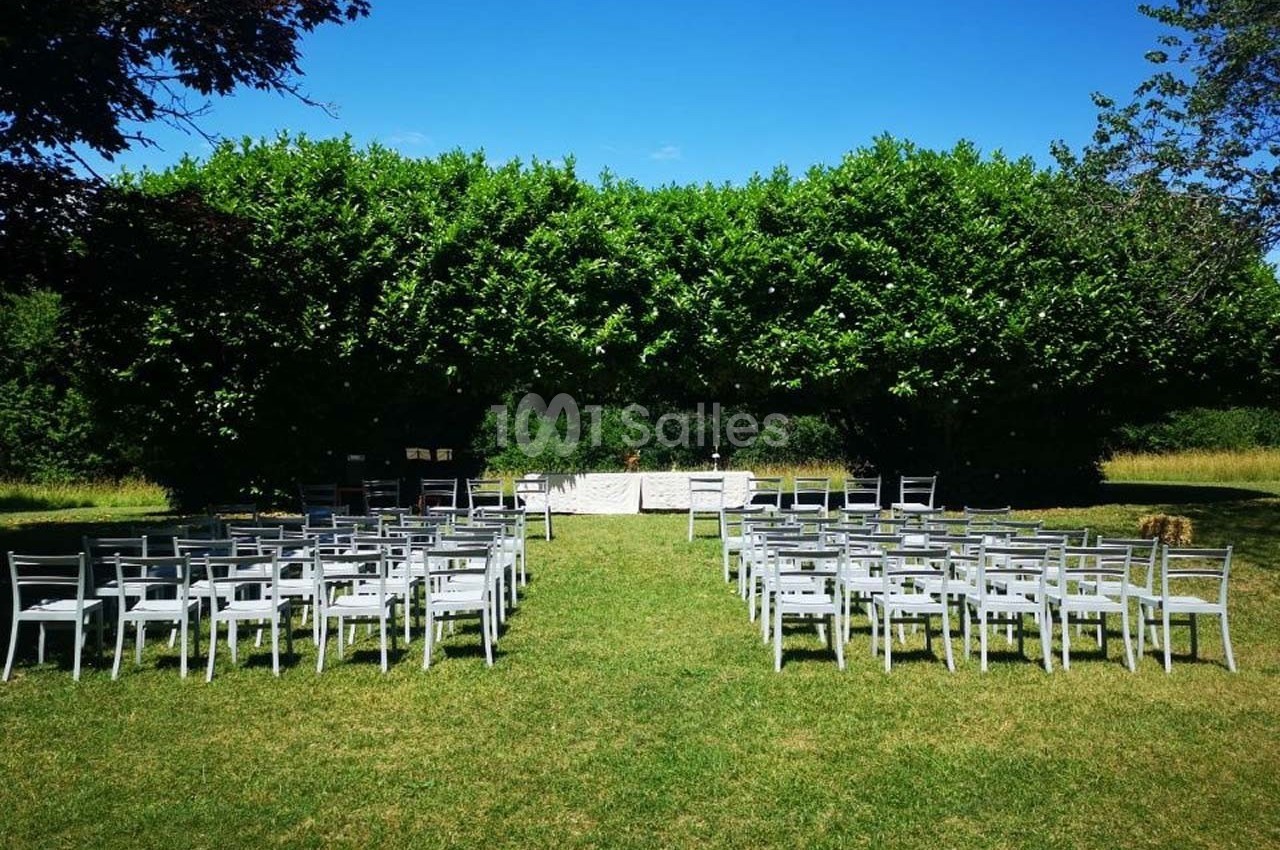 Chaises blanches disposées en rangées sur une pelouse, face à une table décorée, entourées de verdure.