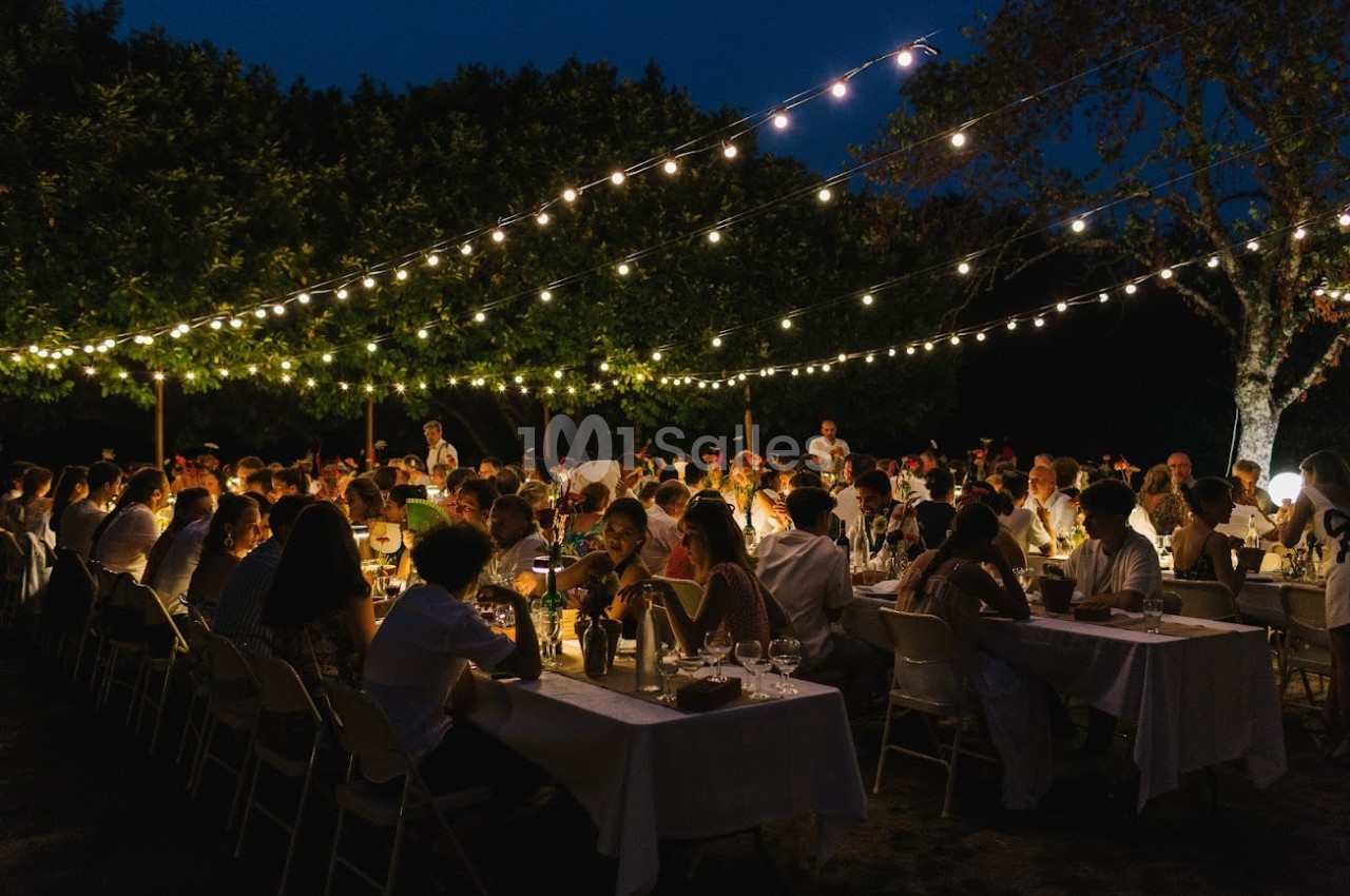 Dîner en plein air sous des guirlandes lumineuses, avec de nombreuses personnes assises à des tables longues.