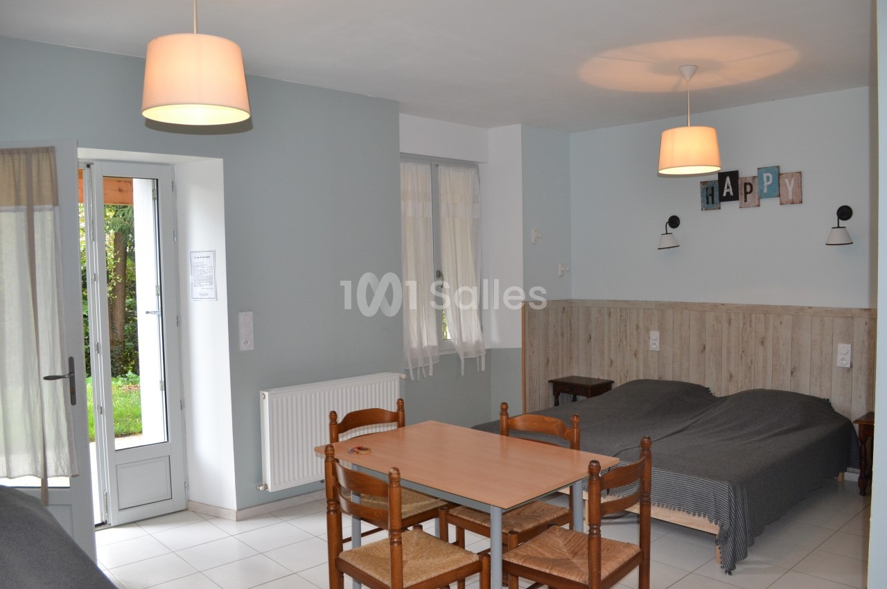 Salle avec deux lits simples, une table en bois et des chaises, éclairée par des lampes suspendues et une porte-fenêtre.