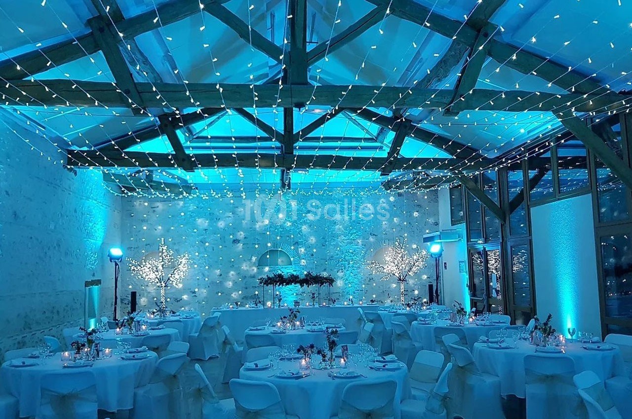 Salle de réception décorée avec des guirlandes lumineuses, tables dressées et ambiance lumineuse bleutée.
