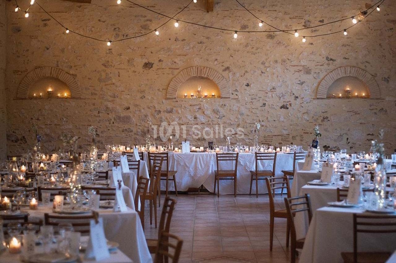 Salle de réception en pierre avec tables dressées, chaises en bois et guirlandes lumineuses suspendues.