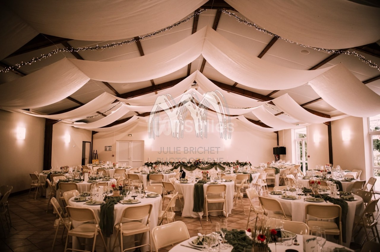Salle de réception décorée avec des tables rondes dressées, nappes blanches et guirlandes lumineuses suspendues.