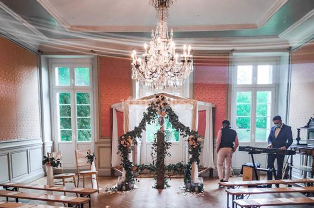 Salle décorée pour une cérémonie avec arche fleurie, chaises alignées et deux personnes préparant l'ambiance musicale.