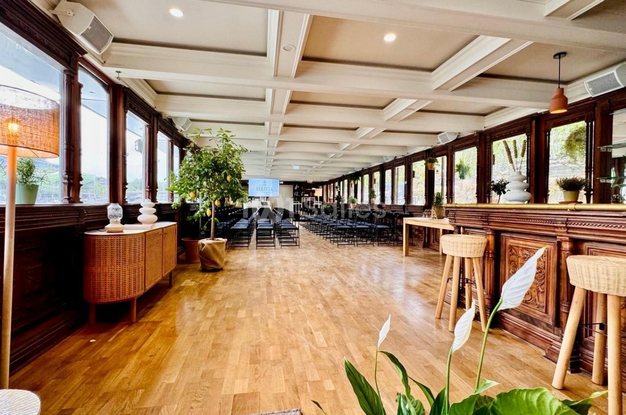 Salle lumineuse avec parquet, bar en bois, plantes décoratives et rangées de chaises disposées face à un écran.