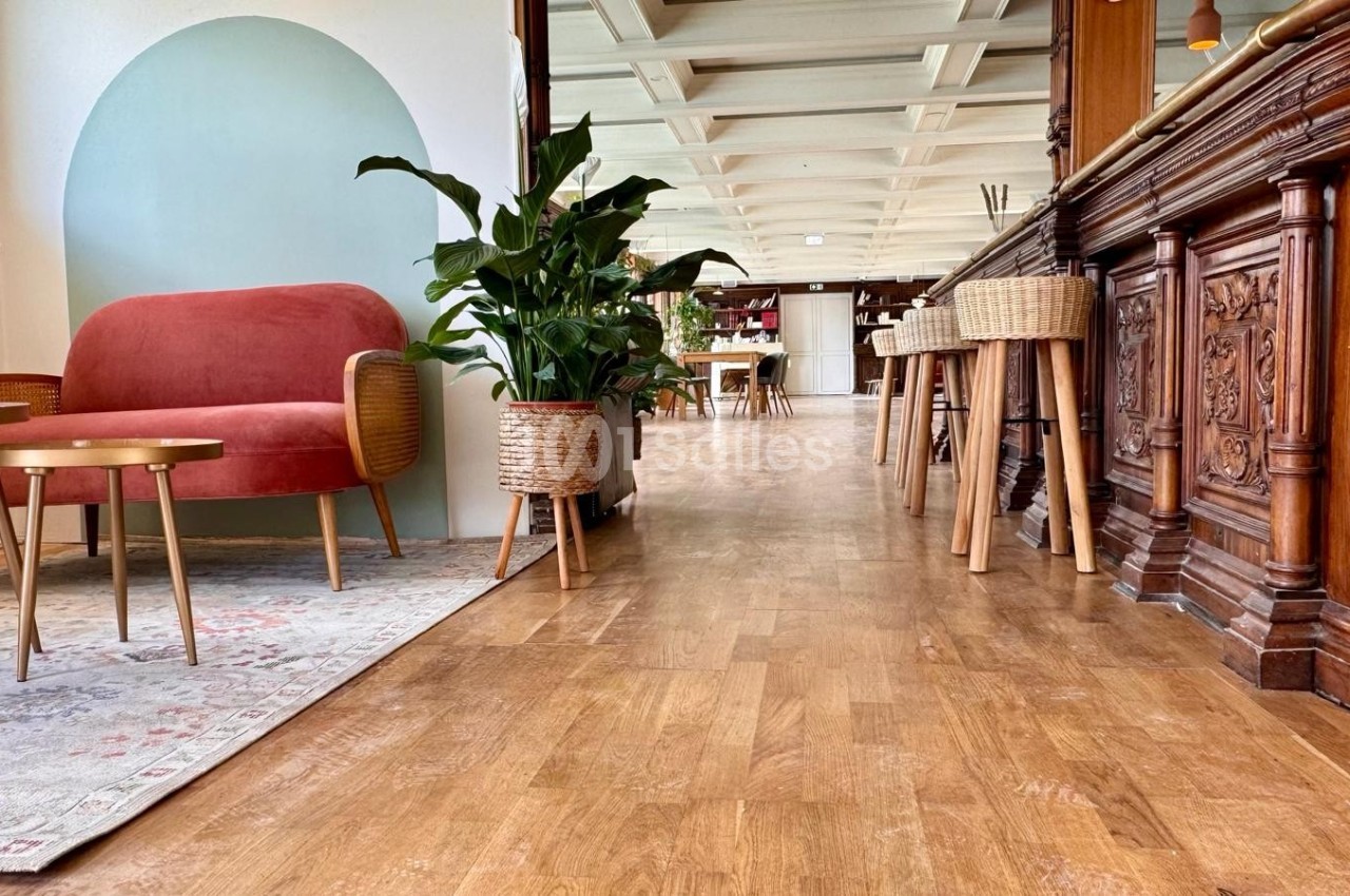 Espace intérieur lumineux avec parquet, fauteuil rouge, tables basses, plantes en pot et comptoir en bois sculpté.