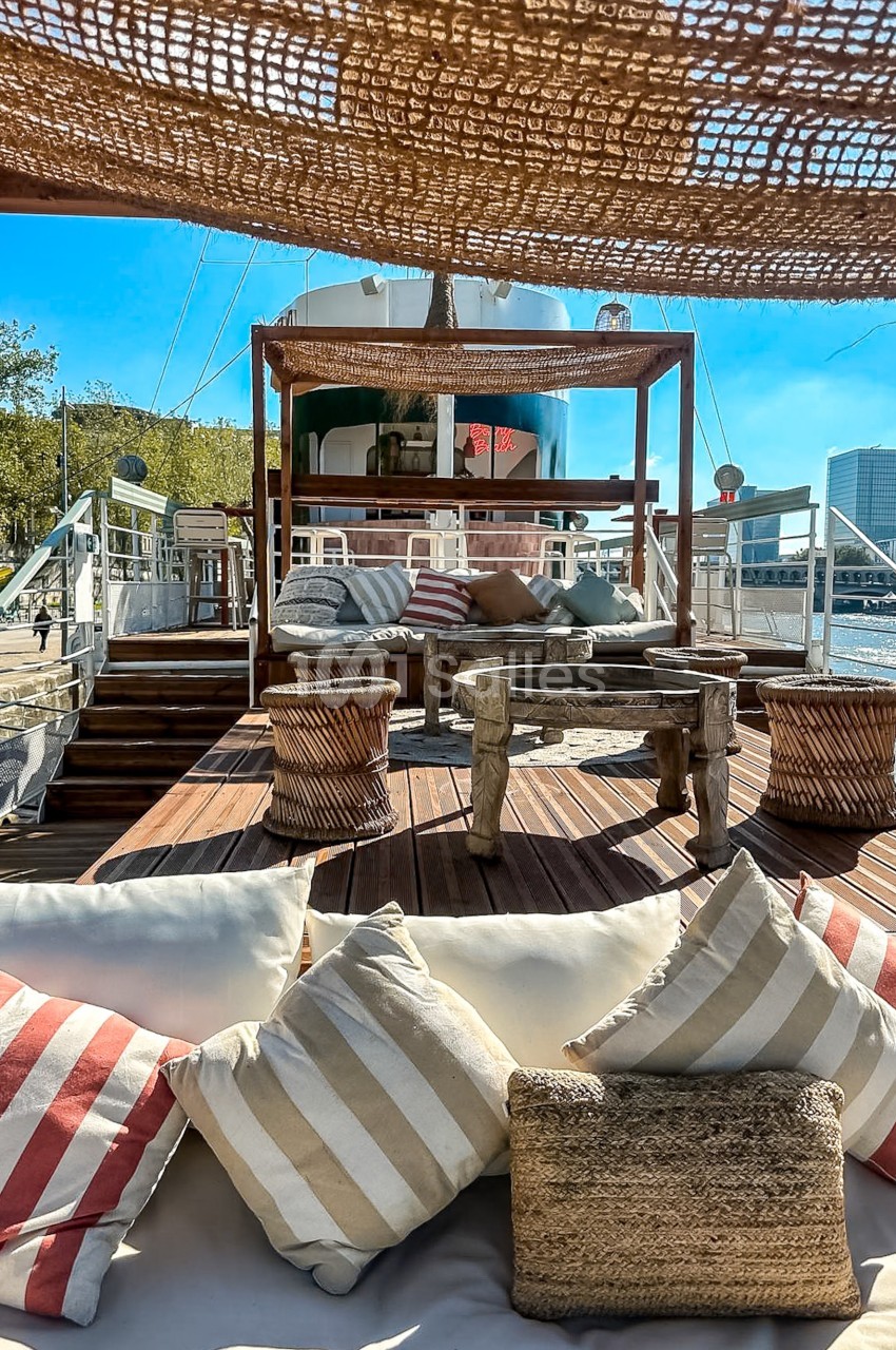 Espace extérieur d'un bateau aménagé avec des coussins, des fauteuils et une pergola en bois, sous un ciel ensoleillé.