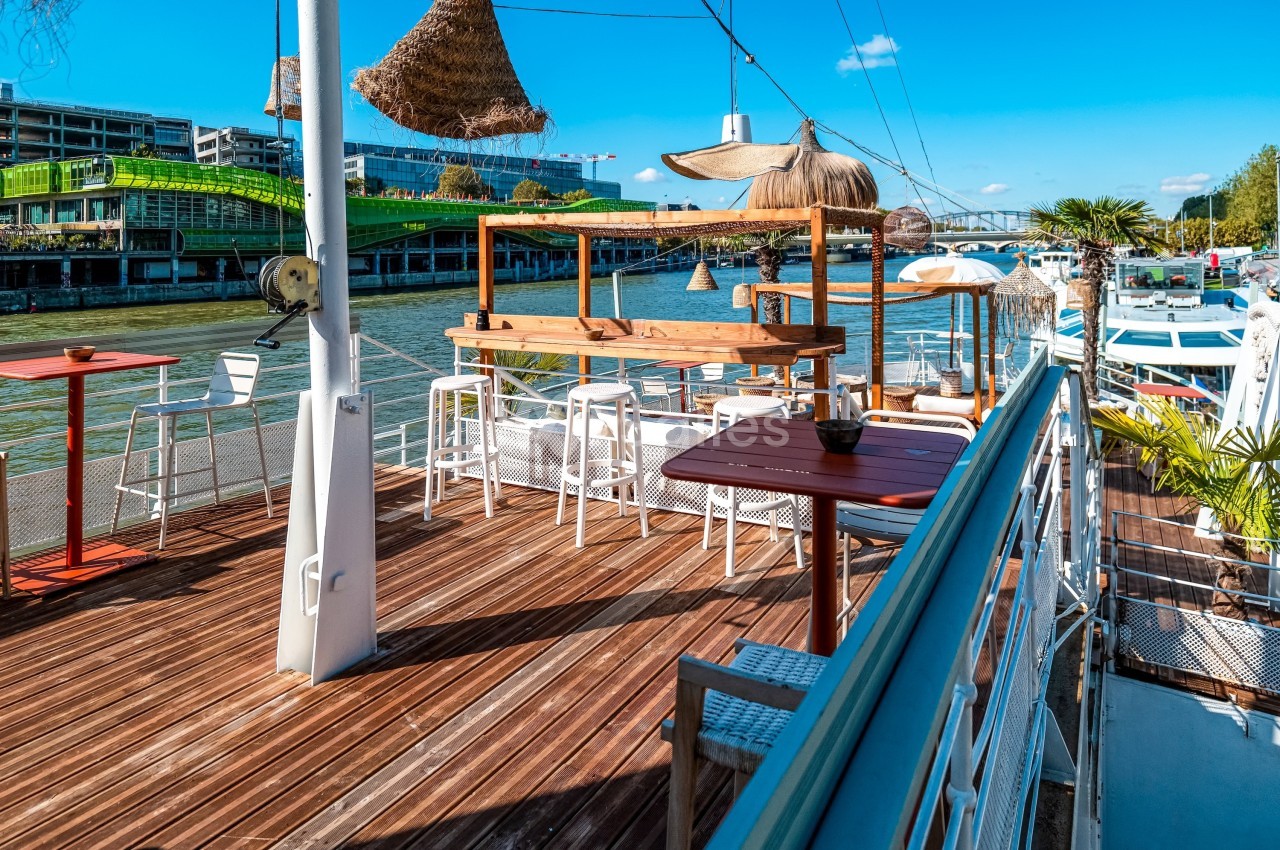 Terrasse en bois aménagée avec tables et chaises, située sur une péniche au bord d'un fleuve par temps ensoleillé.