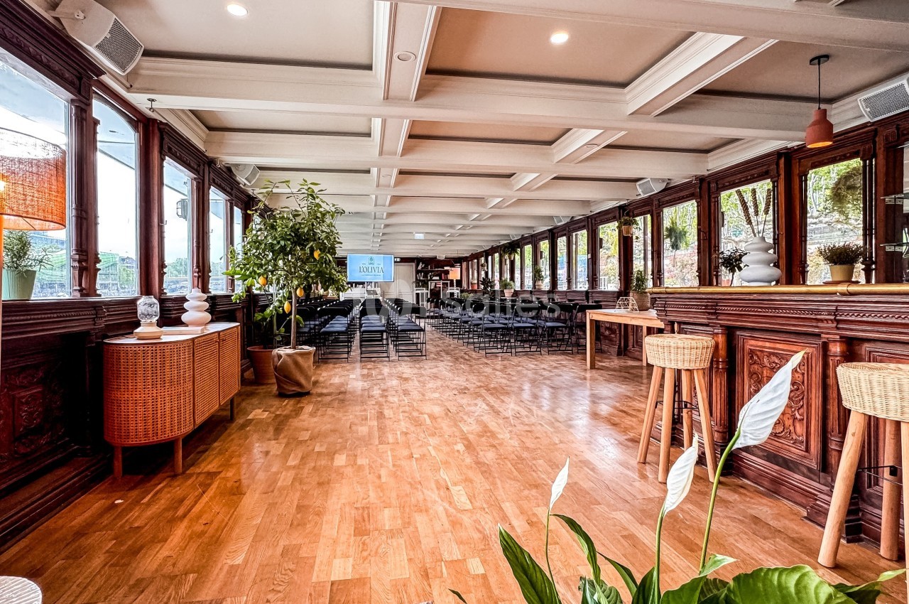 Salle lumineuse avec parquet, grandes fenêtres, plantes décoratives et chaises disposées pour un événement.