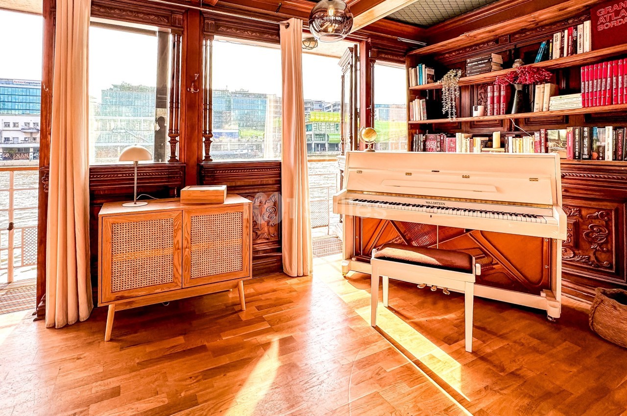 Intérieur lumineux avec piano blanc, bibliothèque en bois, meuble vintage et vue sur l'eau à travers de grandes fenêtres.