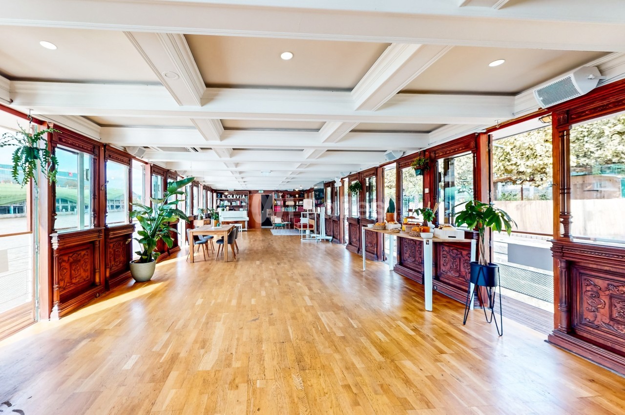 Salle lumineuse avec parquet en bois, grandes fenêtres, plantes et étagères au fond.